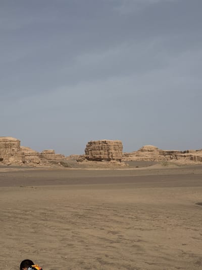 The image depicts a vast, arid desert landscape under an overcast or hazy sky. The photograph appears to be originally a landscape orientation but is presented rotated 90 degrees clockwise.

In the corrected orientation, the upper portion of the frame is dominated by a pale, uniform grey-blue sky, indicating an overcast day or significant atmospheric haze.

Stretching across the mid-ground, below the sky, are distinctive, rugged rock formations. These formations, likely yardangs, are light brown and tan, showing layered and eroded surfaces. They vary in height and shape, creating an irregular, undulating line across the horizon. A narrow, flat strip of ground, possibly a road or pathway, runs parallel to the base of these formations.

The foreground and much of the lower right half of the frame are covered by an expansive sandy or dirt ground. It is light brown with subtle variations in texture, suggesting footprints or wind patterns.

A person is partially visible in the lower right corner of the image (when oriented correctly). Only the back of their head/shoulders are seen. They appear to be wearing a dark hat or have dark hair, and a garment with a prominent pattern, including yellow and possibly other colors.

The lighting is soft and diffused, characteristic of an overcast day, with no harsh shadows present. There is no visible text in the image. The geological features and sandy environment are consistent with desert regions like Dunhuang City, China.