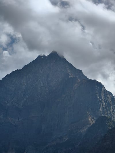 A dramatic, vertical shot captures the imposing, dark, and rugged slopes of a colossal mountain peak. The mountain dominates the frame, its sheer rock face textured with numerous vertical lines and indentations, suggesting a history of erosion and geological formation. The summit of the peak is partially obscured by thick, heavy clouds that hang low, creating an atmosphere of mystery and grandeur. Patches of lighter, scattered clouds drift above and around the main cloud cover, with glimpses of a pale blue sky visible through them. The lighting is subdued, with the dark mountain contrasting sharply against the lighter tones of the clouds. No people, animals, or man-made objects are visible, focusing the attention solely on the raw, natural power of the mountain landscape. The image appears to be a close-up view, emphasizing the scale and imposing nature of the geological formation. This scene is located in the region of Tiling, India.