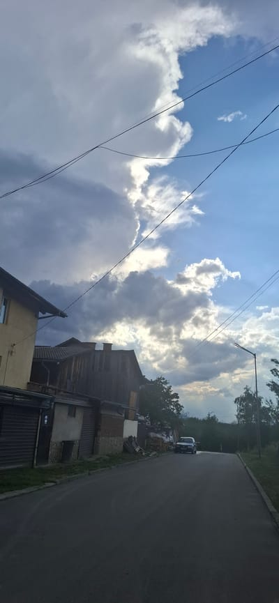 This image, originally rotated, captures a street scene in a rural or semi-rural setting, likely Dobrinishte, Bulgaria. The upper portion of the frame is filled with a dynamic sky, characterized by patches of bright blue interspersed with large, fluffy white cumulus clouds. Bright sunlight breaks through these clouds, illuminating the scene. Several dark power lines or communication cables crisscross the sky.

An asphalt road runs along the right side of the image, gently curving into the distance. Adjacent to the road is a series of buildings, appearing to be a mix of residential homes and outbuildings. These structures display varied architectural elements, including traditional pitched roofs, wooden facades, and walls of concrete, brick, or corrugated metal. In the lower-left foreground, a portion of a light-colored building with a window is visible.

Along the roadside near the buildings, piles of materials, including stacked wood and what seems to be construction debris, are present. A dark-colored car, possibly a sedan, is parked on the shoulder of the road, partly obscured by these materials. A streetlight pole with an extended arm is also visible against the sky. Beyond the immediate buildings and road, dense trees and green vegetation line the horizon.

The time of day appears to be late morning or early afternoon, suggested by the bright sunlight. The weather is partly cloudy, with a mix of blue sky and significant cloud cover. No people or active events are depicted, creating a quiet and still atmosphere. There is no visible text in the image.