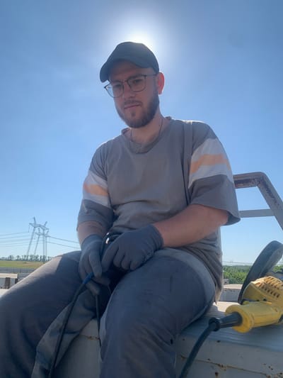 The image features a man in an outdoor setting, with a bright blue sky dominating the background, suggesting it is daytime and likely sunny. He is wearing a grey t-shirt with horizontal stripes of white and orange on the sleeves, grey pants, a dark baseball cap, and clear-rimmed glasses. He also wears grey work gloves and a silver chain necklace.

The man appears to be working on something, as he is holding a tool that resembles a yellow angle grinder with a black cord. His hands, clad in gloves, are manipulating this tool and a cable. He is positioned on what looks like a large, metallic surface, possibly part of industrial equipment.

In the background, power lines and transmission towers are visible, hinting at a location near power infrastructure, which aligns with the provided context of Pivdennoukrainsk, Ukraine, known for its power generation facilities. The overall scene depicts a worker engaged in some form of maintenance or construction, under a clear sky. The sun is very bright, creating a glare in the upper left portion of the image, and illuminating the scene from behind the subject, casting some light on his face. The man's expression is neutral. There is no visible text in the image.