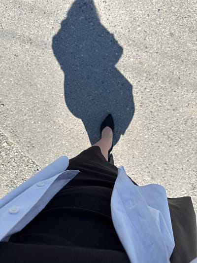 An overhead, ground-level perspective shows a shadow of a person walking on asphalt. The shadow's head is on the left side of the frame, and the body extends towards the right. The person's left foot is visible, wearing a black stiletto heel, and their right leg is obscured by their shadow. They are wearing a black, ribbed skirt and a light blue collared shirt that is open and draped over their shoulders. The asphalt is a light gray, speckled with darker stones and some gravel on the left side. The image does not provide any information to determine the location within Nogir, Russia.