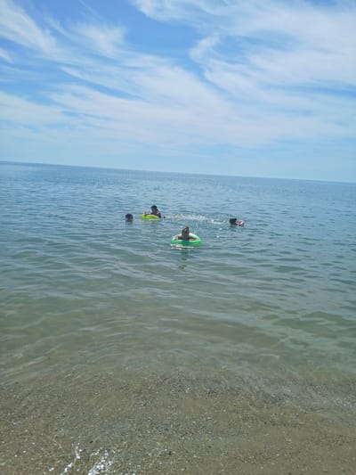 In Kranevo, Bulgaria, four individuals are observed in the calm, clear waters of the Black Sea. The scene is set under a bright, blue sky with scattered, wispy clouds. The shallow, sandy seabed is visible beneath the translucent water, with small pebbles and shells dotting the shore. Two individuals are swimming, while two others are using inflatable tubes. One person is in a green inflatable ring, another in a yellow one. The overall atmosphere is one of leisure and enjoyment in a coastal environment.