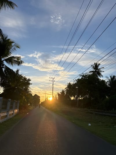 The image depicts a paved road leading towards a setting sun in Geulumpangsulu Timu, Indonesia. The road is lined with lush green vegetation and palm trees on both sides. In the foreground, the road is dark and shows reflections of the sunset. Further down the road, two motorcycles are visible, with riders heading away from the camera. Utility poles with multiple wires stretch across the sky, creating a crisscrossing pattern. The sky is a blend of blue and orange, with scattered clouds illuminated by the setting sun. The overall scene evokes a tranquil, rural atmosphere.