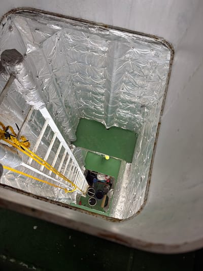 This image depicts a vertical shaft within a structure, likely on a vessel or industrial building. A white metal ladder with yellow ropes attached extends down into the shaft. The walls of the shaft are lined with quilted silver insulation material. At the bottom of the shaft, a green-painted floor is visible, with some objects scattered on it, including dark containers and what appears to be some equipment. The overall impression is one of an internal, utilitarian space. The location in Melaya, Indonesia, suggests this could be part of a port facility, a fishing vessel, or an industrial site within the town. There are no people visible in the image.