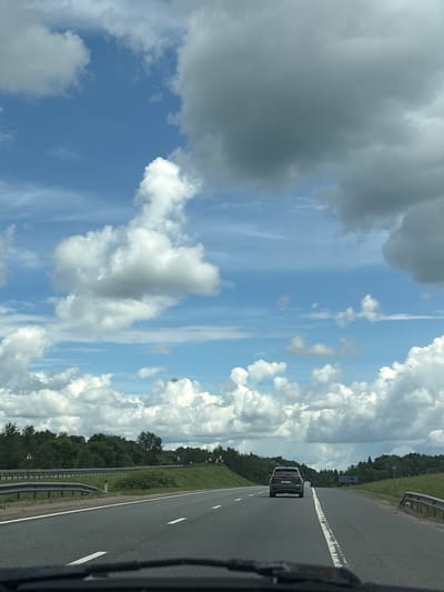 The image shows a highway with a dark SUV driving away from the camera. The road has white lane markings and guardrails. On either side of the road are green trees and grass. The sky is blue with scattered white and gray clouds. The image appears to have been taken from the perspective of another vehicle. There are no visible people in the image. The location is identified as Leshkovo, Russia.