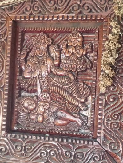 This is a close-up, vertical, eye-level shot of a detailed wooden carving. The carving depicts a central figure with multiple faces, seated on what appears to be a tiger. To the left of this central figure, a smaller figure is depicted lying down, with its head resting on a pillow. To the right of the central figure, another smaller figure with multiple heads is shown in a prayerful pose. The carving is framed by intricate scrollwork. Dried flowers are visible hanging on the right side of the frame, partially obscuring the carving. The wood is dark brown with some reddish-brown accents and lighter highlights from the carving. The lighting is even, and there are no visible shadows. The location in Gojra, India is not discernible from the image.