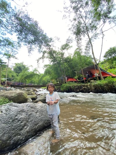 A child stands in a shallow, clear river. She wears a white shirt and light-colored pants, and her feet are immersed in the water. The river flows over rocks and stones. Lush green vegetation and tall trees line the riverbank. In the background, there are camping tents set up in a shaded area. The sky is bright, suggesting a sunny day.