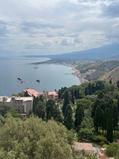 This is an aerial, high-angle shot looking southeast over the city of Taormina, Italy, and the Ionian Sea. The foreground is dominated by lush green trees, including olive trees. In the middle ground, several buildings are visible, including a large pink building with a terracotta roof and a grey, multi-story building with white window frames. A construction crane is positioned between these buildings and the sea.

The sea is calm and a deep blue. Several boats, including what appear to be yachts or ferries, are moored or sailing on the water. The coastline stretches into the distance, lined with a town that extends up the hillsides. The background features a hazy mountain range, likely Mount Etna, under a cloudy sky. The overall impression is of a scenic coastal town nestled between the sea and mountains.