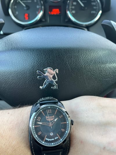 The image depicts a close-up, first-person perspective from inside a vehicle, potentially in Maksimkovo, Russia, though specific geographic identifiers are absent from the visual information. In the foreground, a light-skinned left forearm and wrist are visible, wearing a large, dark-faced Shturmanskie wristwatch with a wide dark leather cuff strap. The watch features a black dial with rose gold-colored Arabic numerals (at 2, 4, 8, 10, 12) and indices for other hours, along with matching hands. A small date window is present at the 6 o'clock position. Text legible on the watch face includes "ШТУРМАНСКИЕ" (SHTURMANSKIE) and "Водозащита 100 м" (Water resistance 100 m).
Behind the wrist, the black, textured leather steering wheel of a Peugeot vehicle is prominent, identified by its central chrome-colored lion emblem. Above the steering wheel, a portion of the vehicle's dashboard instrument cluster is visible. The left circular gauge displays a red illuminated "S". A red, pixelated digital display is situated centrally between the gauges. Paddle shifters are partially discernible behind the steering wheel on both the left and right sides. The scene suggests a stationary or slow-moving vehicle with a driver or passenger present.