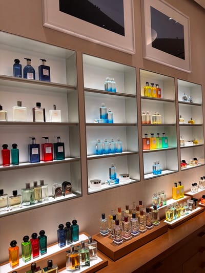 The image displays a well-lit retail display of perfumes. The scene is set within a store, likely a department store or a dedicated brand boutique, with white shelving units showcasing various perfume bottles. The shelving is organized into sections, with multiple rows and columns of products. Some shelves are illuminated with subtle lighting that highlights the bottles.
On the top shelves, there are framed artworks on the wall above the product displays. The artworks appear to be large, possibly photographs, with dark subjects against lighter backgrounds.
The perfumes themselves are presented in a variety of bottle shapes, sizes, and colors. Many are transparent or colored glass, containing liquids that range from clear to vibrant shades of blue, green, red, yellow, and orange. Some bottles have distinct cap designs. There are also a few larger pump bottles and some cylindrical containers that might be lotions or other scented products.
The bottom of the image shows a wooden counter or display surface with more rows of perfumes. These are presented on wooden risers and trays, with some individual bottles and sets. The lighting in this area is also soft and focused on the products.
There are no people visible in the image. The focus is entirely on the organized and aesthetically arranged merchandise. The overall impression is one of a clean, upscale, and professional retail environment. The specific location in Xuanwu District, China is not directly discernible from the visual elements of the image itself.