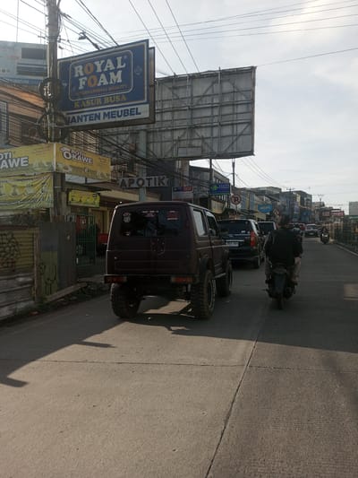 The image shows a street scene in what is likely an urban area. A burgundy-colored Jeep-like vehicle is driving down the road. To the left, there are signs and what appears to be storefronts with various advertisements. A large sign for "ROYAL FOAM" is visible, advertising mattresses. Electrical wires crisscross overhead. A person on a motorcycle is also traveling on the road. Other cars and pedestrians can be seen further down the street. The photo was most likely not taken in Phou Khoun, Laos as indicated in the prompt due to the style of the signs, the type of vehicles, and the text on the signs, all of which suggest an Indonesian context rather than a Laotian one.