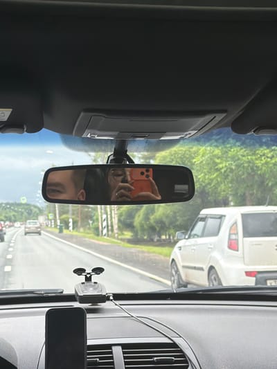 The image is taken from the interior of a car, looking forward through the windshield and up at the rearview mirror. The rearview mirror reflects two people and an orange smartphone. One person, on the left side of the reflection, appears to be a male with a portion of his face visible, including an eye. The other person, on the right side of the reflection, is holding the orange smartphone and appears to be a female wearing glasses. Outside the car, a two-lane road is visible with other vehicles driving in the same direction. The road is flanked by green trees and foliage. In the foreground, on the dashboard, is a black smartphone mounted on a holder, and a radar detector. The location is Mshaga, Russia, as indicated by the prompt. The scene depicts a typical car journey with a self-portrait being taken by one of the passengers.