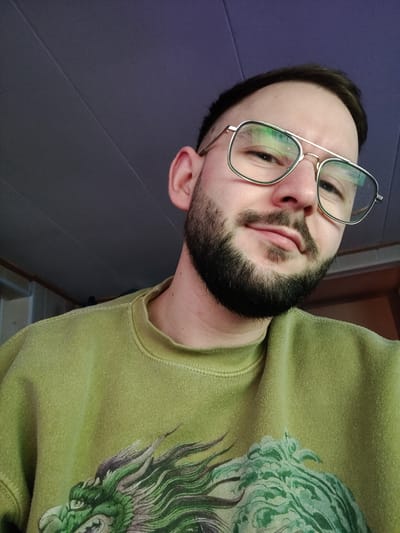 An adult male is depicted in a close-up portrait, taken indoors in the city of Dorst, Netherlands. He has short, dark hair, a full dark beard, and a mustache. He wears square-framed, metallic-rimmed glasses with a double bridge, which show reflections of an unseen light source. He is smiling faintly. He wears a lime green crewneck sweatshirt, which appears slightly worn or pilled, featuring a prominent graphic of a stylized green dragon's head with sharp teeth and flowing details. The background is a dark, out-of-focus paneled ceiling or upper wall, with a subtle purplish hue and linear seams.