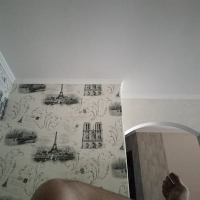 The image shows a bedroom with wallpaper depicting Parisian landmarks such as the Eiffel Tower and Notre Dame Cathedral. The wallpaper is in a grayscale, sketch-like style with flourishes and text fragments. The room has white crown molding along the ceiling. To the right, an arched doorway reveals another room with textured wallpaper. A person's bare foot and lower leg are visible in the foreground, resting on a surface. The scene appears to be indoors, in natural light, suggesting daytime. The overall impression is a home environment, possibly a bedroom, decorated with a travel theme.