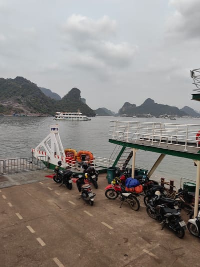 The image depicts a ferry transporting motorcycles in Hải Phòng, Vietnam. Several motorcycles are parked on the ferry deck. In the background, karst formations characteristic of Ha Long Bay are visible, along with other boats on the water. The sky is overcast. Ferry structural elements are visible, including railings, a loading ramp marked "PM15", and life preservers.