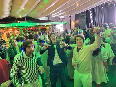 The image captures a lively scene at Foynhagen in Tønsberg, Norway, during what appears to be a silent disco event. The environment is characterized by a large, open tent structure with inflatable white elements and green lighting creating a festive atmosphere. Numerous individuals, mostly young adults, are visible, many wearing headphones, indicating participation in a silent disco. The crowd is engaged in various activities such as dancing, socializing, and holding drinks. The foreground features a man in a black jacket and jeans, holding a drink and gesturing with two fingers. To his left, another man in a striped sweater is also holding a drink and looking towards the camera. The background shows more people, a stage with speakers, and signage, including "Foynhagen" and sponsors like "4human," "DNB," and "CFibico," confirming the location. The overall impression is one of a vibrant, social gathering with a focus on music and entertainment.