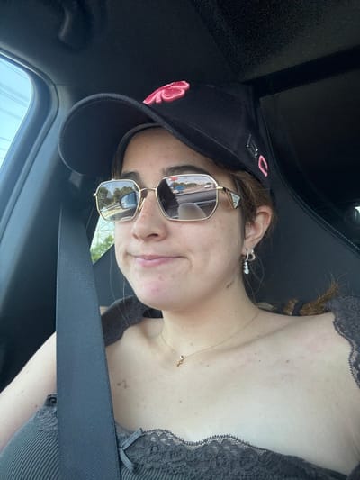 A young adult female is depicted seated in the passenger seat of a vehicle. She wears a black baseball cap featuring a prominent pink four-leaf clover embroidery on the front and a metallic or plastic tag on the side. Gold-framed, hexagonal sunglasses by Prada cover her eyes, reflecting a multi-lane highway scene. This reflection clearly shows other vehicles, a concrete barrier, and green vegetation under a clear sky. Below the sunglasses, her facial expression displays a slight upward curve of the lips. She is dressed in a dark grey ribbed tank top with a lace trim, a thin gold necklace with a small pendant, and silver hoop earrings. A black seatbelt is visible across her chest and left shoulder. The vehicle appears to be in motion on a major thoroughfare, likely the A-1 (Autovía del Norte) highway, within or immediately adjacent to San Sebastián de los Reyes, Spain.