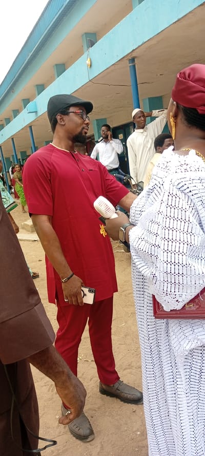The image appears to be taken outdoors in a public space in Goron Dutse, Nigeria. The setting seems to be near a building with a blue and white facade, possibly a school or community center.
The main subjects are a man and a woman engaged in a conversation. The man wears a red tunic and trousers, a black cap, and glasses, holding a white smartphone in his left hand and what appears to be a small electric fan in his right hand. The woman is dressed in a white lace garment with a red head covering and holds a red clutch purse.
In the background, there are several other people. One man in a white shirt is using a cell phone. Another man is wearing a white robe and turban. There are other figures visible in the background, adding to the impression of a gathering or public event.
The ground appears to be sandy or dusty. The scene has the quality of a casual, candid photograph, capturing a moment of interaction between the two central figures amidst the surrounding environment and other individuals.