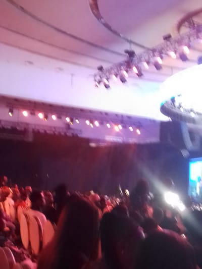 The image shows a crowded indoor event in Satellite Town, Nigeria. A large audience is seated in chairs, facing a stage with lighting rigs overhead. A large screen is visible on the right. The lighting is a mix of white and purple.