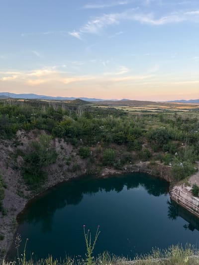 The image captures an overhead view of a deep, dark blue quarry lake surrounded by lush green vegetation and rocky edges. The lake's surface reflects the surrounding trees and the sky. In the foreground, wild grasses and two delicate purple flowers are visible. Beyond the quarry, the landscape unfolds into rolling hills covered in a mix of trees and fields, leading to distant mountains under a pale blue sky with wispy clouds. The scene is bathed in the soft light of what appears to be either dawn or dusk, indicated by the warm hues in the sky. The location is a quarry in Veren, Bulgaria. There are no people or animals visible in the image.