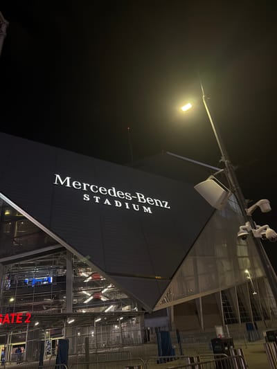 The image captures an exterior view of Mercedes-Benz Stadium in Atlanta, United States of America, at night. The stadium's distinctive angular architecture is prominent, featuring the illuminated "Mercedes-Benz STADIUM" signage in a vertical arrangement. A streetlight casts a bright glow above, and surveillance cameras are mounted on a structure near the stadium's upper section. The right side of the frame reveals elements of the stadium's exterior, including metallic structures, lighting, and signage for "GATE 2" in red. The overall scene suggests an urban environment, with the focus primarily on the stadium's architectural features and lighting.