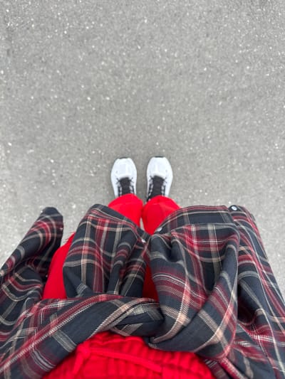 A downward, top-angle shot depicts a person standing on a grey concrete surface. The person is wearing bright red pants and white sneakers with black laces. A dark plaid jacket with red and tan accents is draped around the person's lower body. The setting appears to be outdoors, likely a sidewalk or paved area. The precise location in Zamankul, Russia cannot be determined from the image alone.