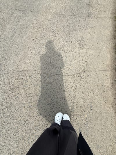 The image provides a high-angle, top-down view of a person's lower body standing on a textured concrete surface. The person is wearing white, perforated slip-on shoes, resembling Crocs, paired with black, wide-leg trousers.

The ground is composed of light gray concrete, densely mixed with small, dark aggregate stones, giving it a coarse texture. A faint, irregular crack runs across the concrete from the upper left towards the center. A prominent, elongated shadow of a person, likely the photographer, stretches across the left and central parts of the frame, indicating bright sunlight and a clear sky. The angle of the shadow suggests it is daytime. Along the very top edge, a narrow strip of dry earth with sparse vegetation or debris is visible, possibly marking the edge of a paved area or sidewalk.

The person appears to be standing still. No specific activity or emotion is discernible. There is no visible text. The location is Aksay, Kazakhstan.