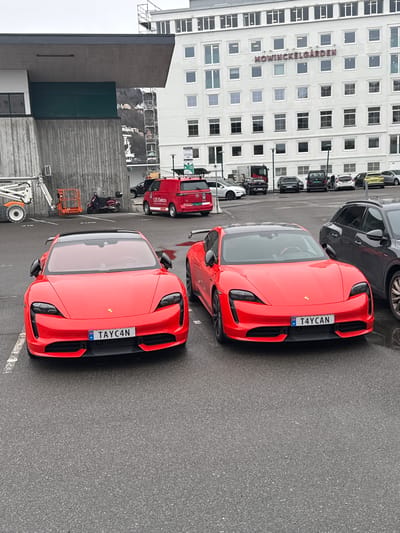 The image shows two red Porsche Taycan vehicles parked in an outdoor parking area. The cars are angled toward the viewer, each displaying a Norwegian license plate with the text "TAYC4N". Behind the cars is a white building identified by signage as "MOWINCKELGÅRDEN", likely a hotel or office building. Other vehicles, including a red Volkswagen van and several dark-colored passenger cars, are visible in the background. A white lift and a small orange trolley are located to the left of the cars. The weather appears overcast. The image is in focus and well-lit, capturing details of the vehicles and the surrounding environment. Given the building's name, the location is likely in the city of Bergen, Norway, where "Mowinckel" is a prominent family name. The scene appears orderly with cars neatly parked. The overall impression is of a modern parking area with a mix of commercial and private vehicles.
