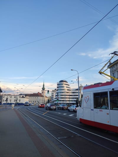 The image captures a view of an urban street scene in Brno, Czechia, with a tram in the foreground. The tram, a modern white and red vehicle, is moving along a set of tram tracks. A large red heart graphic is visible on its side, along with the number "1915".

In the background, several cars are driving on the road. Buildings of various architectural styles line the street, including a church with a green steeple. The sky above is a clear blue with a few wispy clouds. The scene appears to be set in the late afternoon or early evening, as indicated by the lighting. The presence of tram tracks, traffic lights, and street lamps confirms the urban setting. The sidewalk in the foreground is paved with bricks.