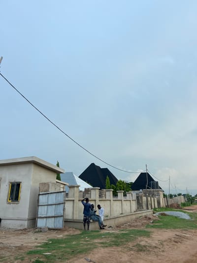 The image depicts an outdoor scene on an overcast day in the city of Zanzi, Nigeria. In the foreground, a dirt path borders a patch of green grass. A wall, constructed from concrete blocks and topped with a plaster finish, runs along the edge of the property. To the left, a light-colored, unfinished building with a yellow-framed window is visible. To the right of the building, a corrugated metal gate is propped open.

Three individuals are present. Two men are seated on the wall near the gate. One man, wearing a white shirt and dark trousers, is looking down at his phone. Another man, dressed in a blue shirt and dark shorts, is standing and appears to be looking towards the right. A third man, also in a white shirt and dark trousers, is seated next to the man with the phone.

Behind the wall, several houses with dark, pyramid-shaped roofs are under construction. Electrical wires are strung across the sky, supported by utility poles. The overall impression is that of a developing neighborhood.