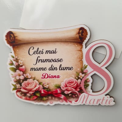This image depicts a decorative item, likely a magnet or ornament, designed for International Women's Day (March 8th). It features a scroll design with text in Romanian. The text reads "Celei mai frumoase mame din lume Diana," which translates to "To the most beautiful mothers in the world Diana." Below this, the date "8 Martie" (March 8th) is prominently displayed as a large number 8 decorated with pink stripes, further reinforcing the occasion. The item is adorned with illustrations of pink roses, smaller white flowers, green leaves, and a pink bow. The background is a plain white surface, consistent with an indoor setting or a product photograph. There is no discernible information within the image that suggests a specific location within the city of Tohatin, Moldova.