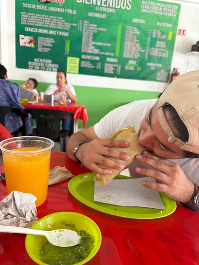 Here is a description of the image:

The photo shows a person wearing a tan baseball cap eating a taco with his hands. There is a green plate with a partially eaten taco, a green plate with a green salsa, and a tall glass of orange juice on a red table.  A crumpled piece of tissue or paper napkin is also on the table.

In the background, other customers are sitting at tables. A menu board with the words "TACOS DE BISTEC, LONGANIZA Y POLLO" is visible. The menu also lists “CERVEZA MICHELADAS Y RUSAS”. Other food options like Quesadillas and Gorditas appear on the menu. The sign also states: "NUESTROS PRODUCTOS SON ELABORADOS AL MOMENTO GRACIAS POR SU PACIENCIA!" and under that in larger font: “TE OFRECEMOS”. The address and telephone number is also on the sign.

The location may be a small taqueria located in San Pedro Cholula, Mexico based on the writing on the sign.