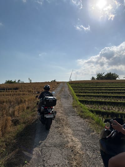 The image shows a sunny day in Selemadeg, Indonesia. Two individuals are on motorcycles traveling along a dirt road. The motorcycle in the foreground is black, and a person's hand holding a phone is visible. The motorcycle behind it is silver and black, with a rider wearing a helmet and a t-shirt. The motorcycle has a black storage box on the back and a license plate that reads "DK5148FUY". To the right of the road are terraced rice paddies, some of which appear to have been harvested, showing dry stalks. To the left of the road is a field of dry grass and some trees. In the background, there are more trees and power lines. The sky is blue with scattered white clouds, and the sun is shining brightly.