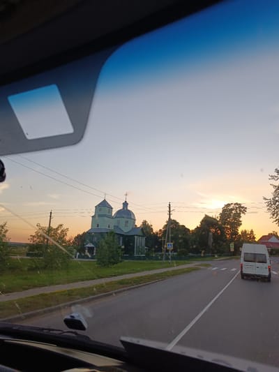 This media file captures a view from inside a vehicle looking out at a rural scene in Porplishchanski Sel'ski Sovet, Belarus, during sunset. The sky exhibits a gradient of blue at the top, transitioning to soft orange and yellow hues near the horizon, indicating the time of day is late evening. Wispy clouds are visible, reflecting the sunset's light.

The primary subject in the mid-ground is a traditional green wooden church with a distinctive onion dome and a cross atop it, silhouetted against the sky. The church appears to be made of dark green wood and has several windows. Adjacent to the church are scattered trees with lush green foliage and a few power lines stretching across the frame.

In the foreground, the dashboard and windshield of the vehicle are visible, with a dashcam mounted on the windshield. A white minivan is seen driving away from the camera on a paved road that runs through the scene. The road has clear white lane markings and a pedestrian crossing. Yellow traffic signs, including a pedestrian crossing sign, are visible along the roadside.

The overall setting is a peaceful, rural landscape with a church as a prominent landmark, bathed in the warm light of the setting sun. The presence of the road and the minivan suggests a journey or travel. The weather appears clear and calm, typical of a summer evening. No people are directly visible.

The visible text on the back of the white minivan is a license plate that reads "E 5906 FE-7".