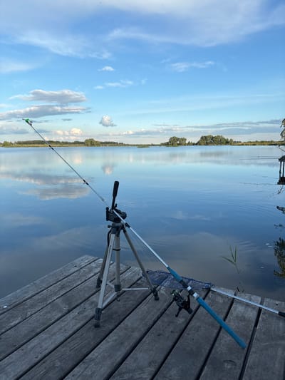 The image depicts a tranquil scene at Khryashchevka, Russia, specifically a view over water, likely a lake or river. A wooden pier or platform dominates the foreground. Resting on the pier are two fishing rods, one secured on a tripod. A fishing net also lies on the pier next to the rod and tripod. The water reflects the blue sky and clouds, suggesting a calm, clear day. In the distance, a line of trees marks the opposite shore. The sky is blue with white clouds.