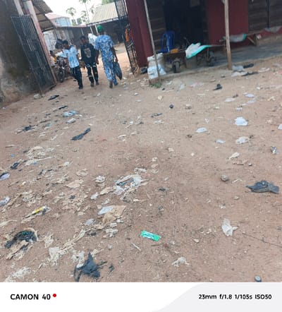 An open area features a dirt ground strewn with plastic debris and trash. Three people walk away from the camera near buildings with corrugated metal. One person carries a dark bag. A motorcycle stands nearby. A wheelbarrow and other items sit outside a structure with a red support beam. The scene shows signs of litter and disrepair.