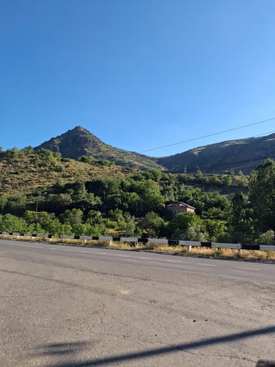 This is a landscape photograph taken on a clear, sunny day, likely in the mid-morning or early afternoon, given the bright sunlight and the shadows. The image features a mountainous scene with a road running alongside. The mountain on the left is steep and covered in vegetation, with a prominent peak. Lush green trees grow densely on the lower slopes and extend towards the right side of the frame. A single-story house with a reddish-brown roof is visible nestled amongst the trees on the hillside. A power line runs across the scene, with utility poles also visible.
On the right side of the image, a paved road occupies a significant portion of the frame. The road appears to be in decent condition with some cracks and visible wear. A striped guardrail, alternating black and white, separates the road from the vegetated area. The overall impression is one of a rural, mountainous setting. There is no visible text in the image.