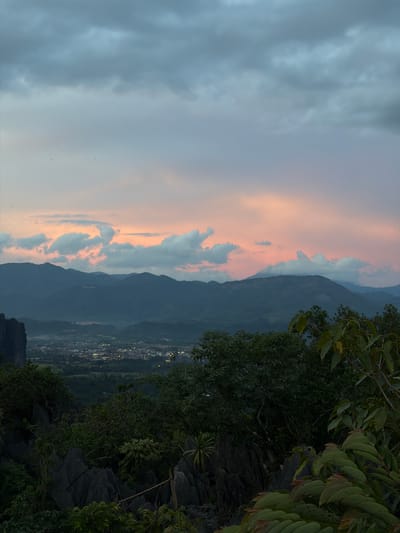 This image captures a scenic view of Vang Vieng, Laos, at dusk. The scene is dominated by rolling, dark blue mountains that recede into the distance. In the sky, the fading light of sunset paints the clouds in soft hues of orange and pink, contrasting with the muted grey of the overcast sky.

In the foreground, lush green foliage and rocky terrain frame the view. To the right, a cluster of trees with broad leaves creates a dense, shadowy foreground. Below and to the left of the trees, a town or village is visible in the valley, its lights twinkling like scattered jewels against the darkening landscape. The lights suggest that night is beginning to fall, and the weather appears to be cloudy or overcast.

The perspective is from a high vantage point, looking down and across the valley. The overall mood is tranquil and serene, with a sense of natural beauty and the gentle transition from day to night. There are no people or explicit objects of human creation other than the distant village lights, emphasizing the natural landscape.