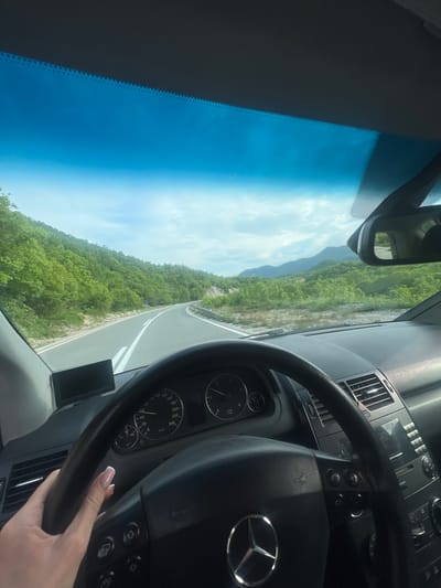 The image depicts a view from inside a car, likely a Mercedes-Benz, as suggested by the steering wheel logo and dashboard design. The car is driving on a winding road through a landscape filled with lush greenery. Mountains can be seen in the distance, and the sky is a clear blue with some clouds. A hand with manicured nails is visible on the steering wheel, implying someone is driving. The dashboard and speedometer are partially visible, providing a sense of the car's interior. The image is possibly taken in Očinići, Montenegro, given the mention of the location.
