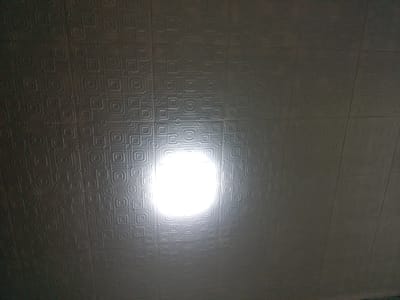The image shows a close-up of a textured ceiling with a repeating geometric pattern of concentric squares. A bright light source is visible in the center of the frame, casting a strong glare and obscuring some of the detail in the pattern directly around it. The ceiling appears to be made of tiles or panels. The overall lighting suggests an indoor environment, possibly a room or hallway. There are no people or other objects depicted in the image. The location in Asino, Russia, is not discernible from the visual information provided in the image.