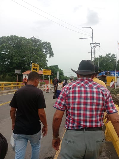 The image depicts a group of people walking on a street in Ureña, Venezuela. In the foreground, two men are walking away from the camera. The man on the left is wearing a black t-shirt and light blue jeans. The man on the right is wearing a black cowboy hat, a red, white, and blue plaid shirt, and grey trousers. They are walking on a street with a yellow barrier on the right side. In the background, there is a crowd of people walking, along with various signs, utility poles, and trees. The weather appears overcast. A 'DESVIO' (Detour) sign can be seen, indicating possible roadwork ahead.