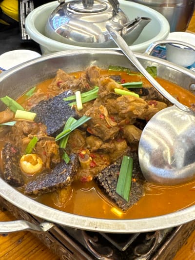 The image depicts an elevated view of a metal cooking pot brimming with a simmering stew, positioned on a dark, ornate metal stand on a light brown wooden table. The stew comprises irregular chunks of brown meat, dark, spongy, textured pieces (resembling blood tofu), and segments of green scallions, all immersed in a thick, orange-brown broth seasoned with red chili flakes. A single yellowish, ovoid object is visible within the stew on the left side. A large, reflective metal ladle is partially submerged on the right. In the blurred background, a light-colored basin holds a shiny metal teapot, positioned next to a smaller metal container and a white ceramic bowl with a blue rim. No individuals are present. The scene represents a prepared meal in Fenghuang, China.
