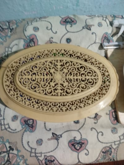 This is a close-up, overhead shot of an oval-shaped, carved, cream-colored decorative object placed on a light-colored fabric with a floral pattern. The fabric has pink, blue, and black floral motifs and thin black lines forming a decorative pattern. The decorative object has an ornate, intricate openwork design with swirling patterns. The background of the image is a light blue wall. The object appears to be made of a material like bone or ivory. There are no people or actions depicted in the image. The scene appears to be indoors. The location in the city of Ananyevo, Kyrgyzstan is not discernible from the image itself.