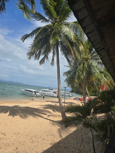 The image depicts a beach scene in Ban Khlong Hia, Thailand. Several speedboats are pulled up onto the tan-colored sand, partially submerged in the shallow water. Two people are near the closest boat, which is labeled "Diamond Sea." Palm trees are prominent, framing the scene, casting shadows on the sand. A roof is visible in the upper left corner. The sky is partly cloudy. Red-and-white umbrellas and additional vegetation are visible on the shore.