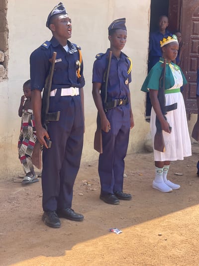 The image captures an outdoor scene in Andaha, Nigeria, depicting a formal gathering or parade involving several young individuals in uniform. The bright daylight and distinct shadows suggest it is daytime, with an unpaved, dusty ground serving as the setting.
In the foreground, two young males stand in formation, seemingly at attention. Both are dressed in navy blue uniforms with gold-colored epaulets, white belts, and matching garrison caps. They each hold a wooden mock rifle. The male closest to the viewer has a nametag on his uniform that reads "AWNIKI OIG". Behind the second male, a young child in a patterned outfit is partially visible.
Adjacent to them, a young female also stands formally. She wears a green and white uniform, comprising a green top with white trim, a white skirt, and a white headpiece with yellow geometric patterns. She has white socks adorned with green text that reads "ARISA IS" (the last word partially obscured) and white athletic shoes. She also holds a wooden mock rifle.
In the background, several other individuals, including children, are visible observing the scene from a doorway of a light-colored building with a dark wooden door. The disciplined postures of the uniformed individuals suggest they are part of a youth organization or cadet corps participating in a drill or ceremonial event.