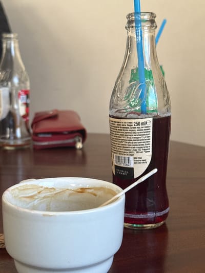 The image shows a table with various objects. There's a half-empty Coca-Cola bottle with a blue straw sticking out of it. Next to it is an empty, white coffee mug with coffee stains inside. A red wallet is also present, slightly out of focus in the background. A second, empty bottle is barely visible in the bottom left corner. The setting appears to be indoors, possibly in a café or restaurant. The Coca-Cola bottle label indicates it is a Bulgarian product, with the contact information being in Sofia, Bulgaria, but does not indicate the location of Пампорово. The image lacks any information that connects it to the city of Пампорово.