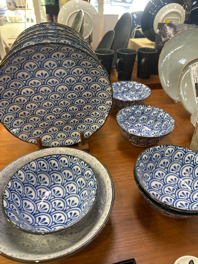 This is a medium-shot, indoor, eye-level photograph of a display of blue and white patterned ceramic tableware on a wooden surface. The most prominent items are a stack of large plates with a repeating fan or scale-like pattern, and several smaller bowls with the same pattern. In the background, there are other ceramic items, including mugs and larger plates, some of which are out of focus. The lighting is neutral, and the overall impression is of a retail display. There are no people visible in the image. The image does not contain any information that would allow its location in Bellingen, Australia to be determined.