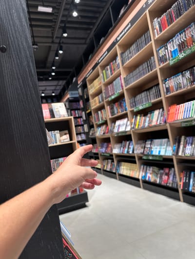A first-person perspective shot captures a hand reaching out towards bookshelves in what appears to be a store or library. The bookshelves, made of light wood, are filled with various items, some of which are identifiable as books and possibly DVDs or video games due to their spines. The items are neatly arranged on multiple shelves, with some displaying promotional signs, including one showing "50%". The environment suggests an indoor retail or educational setting with artificial lighting from overhead fixtures. The foreground is dominated by a dark, textured surface, possibly a pillar or display unit, and the arm and hand of the person taking the photo. The overall impression is one of browsing or selecting items from a well-stocked collection. No specific location in Глебовское, Russia can be identified from the visual information provided.