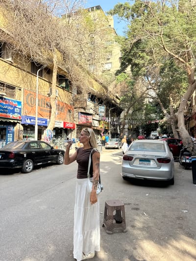 A photograph shows a street scene in Cairo, Egypt. A woman stands in the street, drinking from a plastic bottle. She wears a brown top and a long white skirt. A black car is parked on the street near her. Behind the car, store signs are visible in Arabic and English, including "ENGINEERED," "MICHELIN," and "petlas." Above the stores, the buildings appear weathered, and trees line the street. A silver car and a small brown stool are also on the street. Decorations are hanging along the street.
