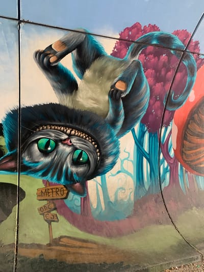 This media file displays a detailed, vibrant mural painted on what appears to be tiled ground, possibly a sidewalk or public square. The central subject is a depiction of the Cheshire Cat from Alice in Wonderland, rendered in a stylized, slightly menacing manner. The cat is predominantly black and blue, with striking emerald green eyes and a wide, toothy grin. Its body is contorted, with paws visible and a long tail curving upwards.
Behind the cat, the scene is a surreal, dreamlike landscape. A large, reddish-brown object, possibly a giant mushroom or a whimsical fruit, occupies the upper portion of the image. Stylized trees with purple foliage and abstract blue branches fill the background, creating a fantastical atmosphere.
In the lower right quadrant, a small wooden signpost is visible. One sign reads "METRO" in vertical letters, while two smaller, angled signs below it appear to have illegible markings, possibly "ODC" and "CS". These signs, along with the known location context of Odivelas, Portugal, suggest the mural is located near a metro station or in a public area within the municipality.
The image is captured at a close-up perspective, tilted to the side, giving a sense of the scale and placement of the artwork within its environment. The tiling technique and the slight wear on the surface suggest it is an outdoor installation. The lighting appears to be natural daylight, though the exact time of day is not discernible. The overall mood is whimsical and slightly unsettling, characteristic of the Cheshire Cat's persona and the Wonderland setting.